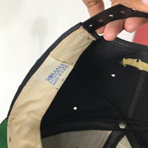 May include: Black baseball cap with a green brim. The interior features a white lining and a tag that reads "ROMAN RANDOLPH, MA MADE IN U.S.A." The cap has a black adjustable strap.