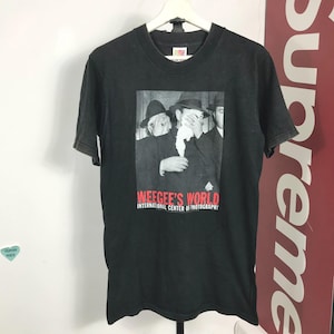 May include: Black t-shirt featuring a black and white photograph of three men in hats, one covering his face with a white cloth. The text "WEEGEE'S WORLD INTERNATIONAL CENTER OF PHOTOGRAPHY" is printed in red below the image. The shirt is hanging on a white hanger.