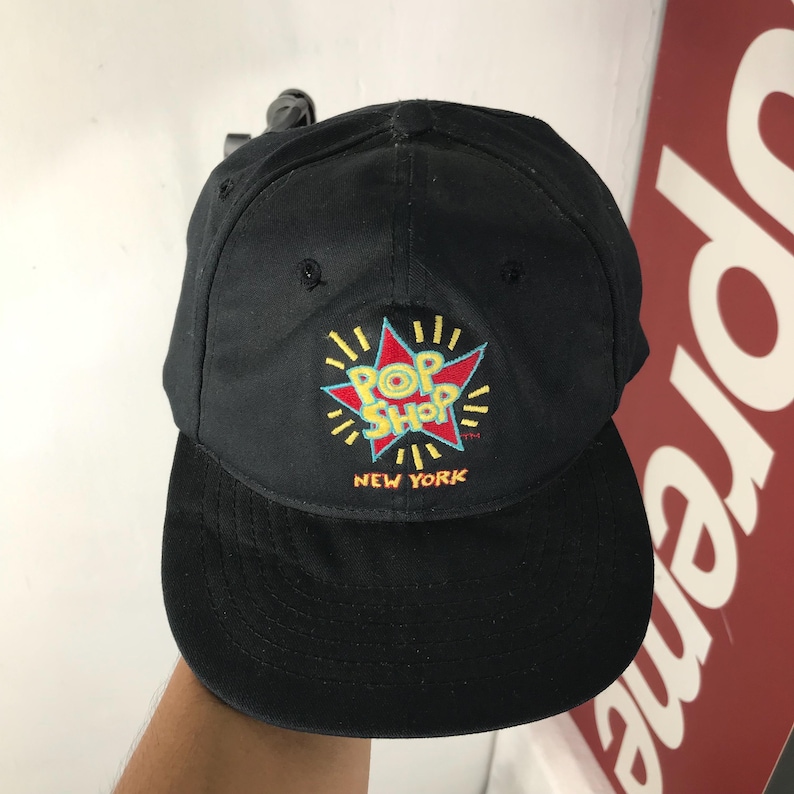 May include: Black baseball cap with a flat brim. The cap features a colorful embroidered logo that reads "POP SHOP NEW YORK" in a starburst design. The hat is made of a durable fabric and has a classic, casual style.