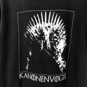 Może przedstawiać: Czarny t-shirt z białą grafiką przedstawiającą osobę na tronie, z koroną u g&oacute;ry. Tekst "KANONENVOGEL" jest wydrukowany na dole na biało. Projekt jest zamknięty w białej prostokątnej ramce.