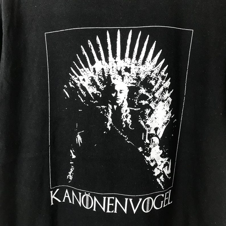 Może przedstawiać: Czarny t-shirt z białą grafiką przedstawiającą osobę na tronie, z koroną u g&oacute;ry. Tekst "KANONENVOGEL" jest wydrukowany na dole na biało. Projekt jest zamknięty w białej prostokątnej ramce.