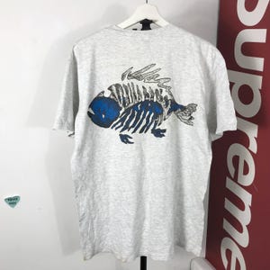 Może przedstawiać: Jasnoszara koszulka z niebiesko-czarnym wzorem szkieletu ryby na plecach. Grafika ryby ma szczegółową strukturę szkieletu z niebieskimi akcentami i stylizowanym wyglądem. Koszulka wisi na białym wieszaku.
