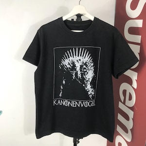 Może przedstawiać: Czarny t-shirt z białą grafiką przypominającą koronę w białym prostokącie. Tekst "KANONENVOGEL" jest wydrukowany na dole na biało. Koszulka wisi na białym wieszaku.