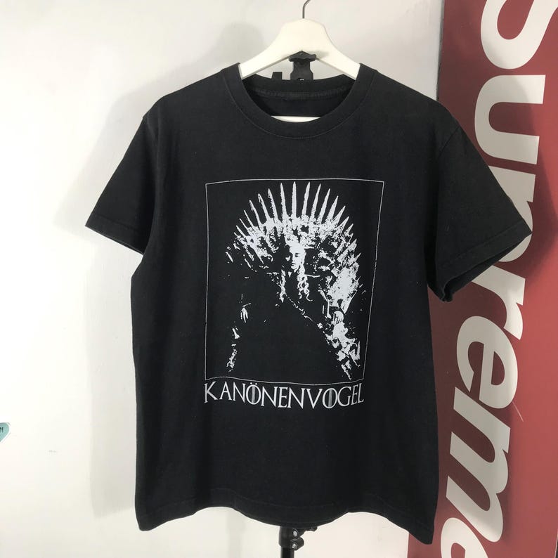Może przedstawiać: Czarny t-shirt z białą grafiką przypominającą koronę w białym prostokącie. Tekst "KANONENVOGEL" jest wydrukowany na dole na biało. Koszulka wisi na białym wieszaku.