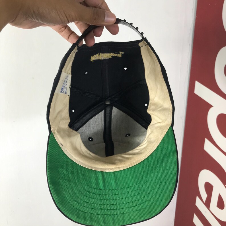 May include: Black baseball cap with a green brim and a beige interior. The cap has a black strap and a tag on the inside. The cap is held up by a hand.