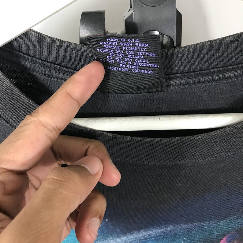 Puede incluir: Primer plano de una camiseta gris oscuro con una etiqueta. La etiqueta dice "MADE IN U.S.A." e incluye instrucciones de lavado. La camiseta est&aacute; sujeta por una mano.