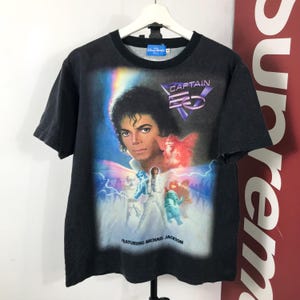 May include: Black t-shirt featuring Michael Jackson with the text "CAPTAIN EO" and "FEATURING MICHAEL JACKSON". The graphic includes images of characters from the film. The shirt is hanging on a white hanger.