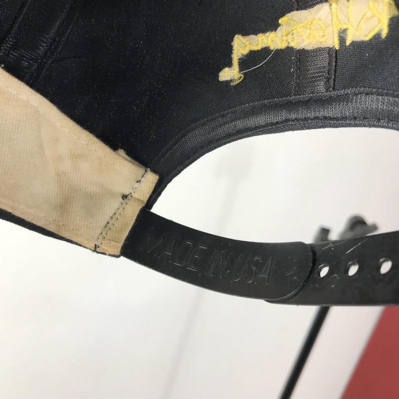 May include: Black baseball cap with a curved brim and adjustable strap. The strap is embossed with "MADE IN USA". The underside of the brim is beige. The cap has a yellow design on the top.