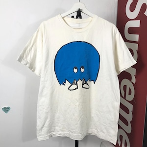 May include: Off-white t-shirt featuring a blue cartoon character with eyes and feet. The graphic is centered on the front of the shirt. The shirt is hanging on a white hanger.