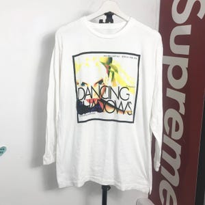 May include: White long-sleeve shirt featuring a graphic from the "Dancing Shadows" album. The graphic is a colorful image of a person's face with the album title in bold letters. The shirt is hanging on a white hanger.