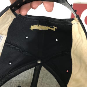 May include: Black baseball cap with a snapback closure. The underside of the cap is black with a beige sweatband and a few small ventilation holes. The label reads "MADE IN U.S.A."