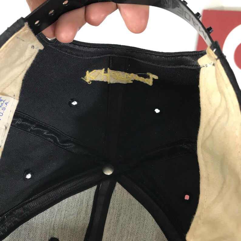 May include: Black baseball cap with a snapback closure. The underside of the cap is black with a beige sweatband and a few small ventilation holes. The label reads "MADE IN U.S.A."