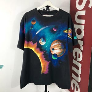 Puede incluir: Camiseta negra con un dise&ntilde;o colorido de tem&aacute;tica espacial. El gr&aacute;fico muestra planetas, un cometa y un sol brillante sobre un fondo azul oscuro y negro. La camiseta tiene mangas cortas y cuello redondo.