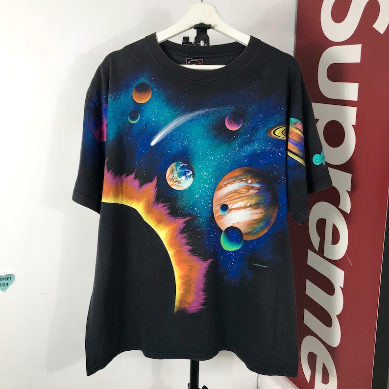 Puede incluir: Camiseta negra con un dise&ntilde;o colorido de tem&aacute;tica espacial. El gr&aacute;fico muestra planetas, un cometa y un sol brillante sobre un fondo azul oscuro y negro. La camiseta tiene mangas cortas y cuello redondo.