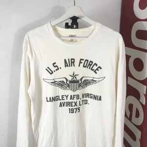 May include: Off-white long-sleeved shirt featuring the text "U.S. Air Force" in black, a winged emblem, and the text "Langley AFB, Virginia, Avirex Ltd., 1975." The shirt is displayed on a white hanger.