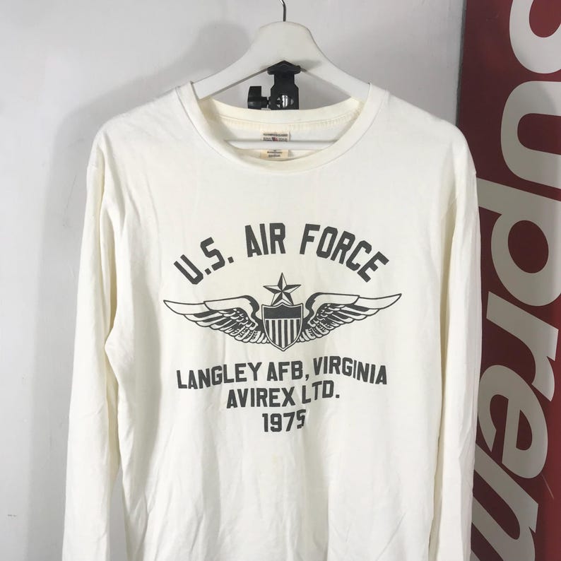 May include: Off-white long-sleeved shirt featuring the text "U.S. Air Force" in black, a winged emblem, and the text "Langley AFB, Virginia, Avirex Ltd., 1975." The shirt is displayed on a white hanger.