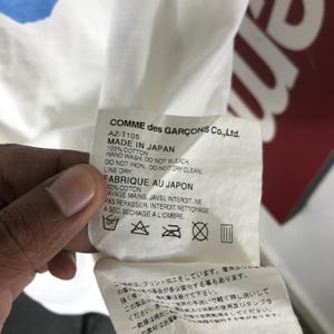 May include: A white fabric tag with black text from Comme des Gar&ccedil;ons Co., Ltd. The tag includes washing instructions and the text "MADE IN JAPAN." The tag is held by a person.