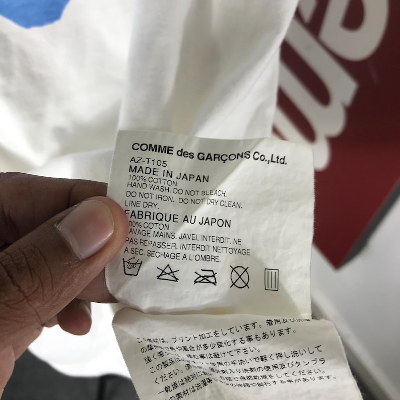 May include: A white fabric tag with black text from Comme des Gar&ccedil;ons Co., Ltd. The tag includes washing instructions and the text "MADE IN JAPAN." The tag is held by a person.