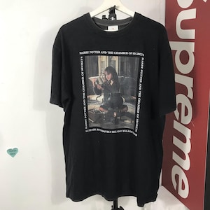 May include: Black t-shirt featuring a graphic from "Harry Potter and the Chamber of Secrets." The image shows a scene from the movie with text around the border. The shirt is hanging on a white hanger.