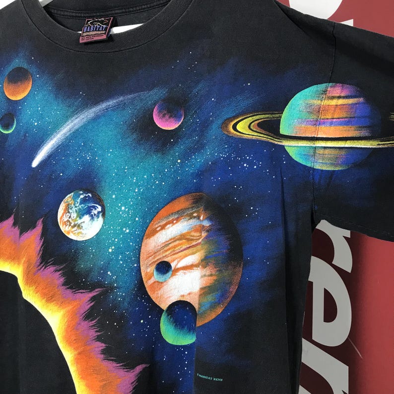 Puede incluir: Camiseta negra con un vibrante dise&ntilde;o tem&aacute;tico espacial. El dise&ntilde;o incluye planetas coloridos, un cometa y un fondo estrellado. La camiseta tiene cuello redondo y mangas cortas. La marca "Habitat" es visible en el cuello.