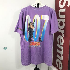 May include: Purple t-shirt featuring a cartoon monkey in a tuxedo holding a gun, with the number "007" in white and blue. The shirt also has the text "LCW KIKIKI" and "Secret Agent".