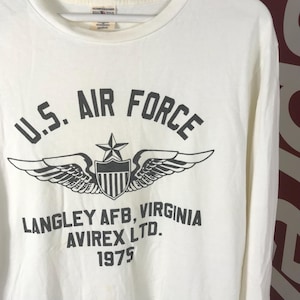 May include: Off-white long-sleeved shirt featuring the text "U.S. AIR FORCE" above a winged emblem and shield. The shirt also displays "LANGLEY AFB, VIRGINIA", "AVIREX LTD.", and the year "1975" in black lettering.