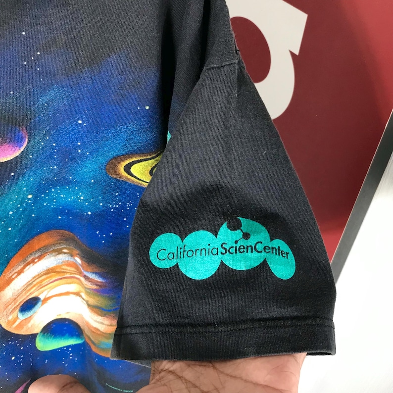 Puede incluir: Camiseta negra con un estampado colorido de tem&aacute;tica espacial con planetas y estrellas. La manga muestra el texto "California Science Center" en color turquesa. La camiseta es de material suave.