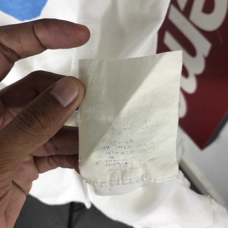 May include: A close-up of a white fabric tag with printed text. The tag is attached to a white garment. The background includes a red and white logo.