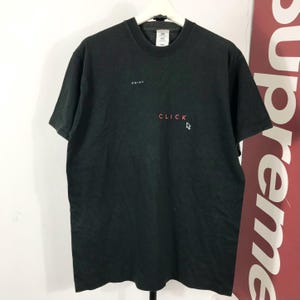 Op de afbeelding: Een zwart t-shirt met korte mouwen voorzien van het woord POINT in kleine witte letters, het woord CLICK in rode letters en een wit computermuiscursor-pictogram rechts van de tekst op de voorzijde van het kledingstuk.