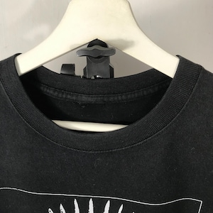 Może przedstawiać: Czarny t-shirt z okrągłym dekoltem i białym wzorem graficznym na dole. Koszulka wisi na białym wieszaku z czarnym zaciskiem. T-shirt jest wykonany z miękkiego materiału.