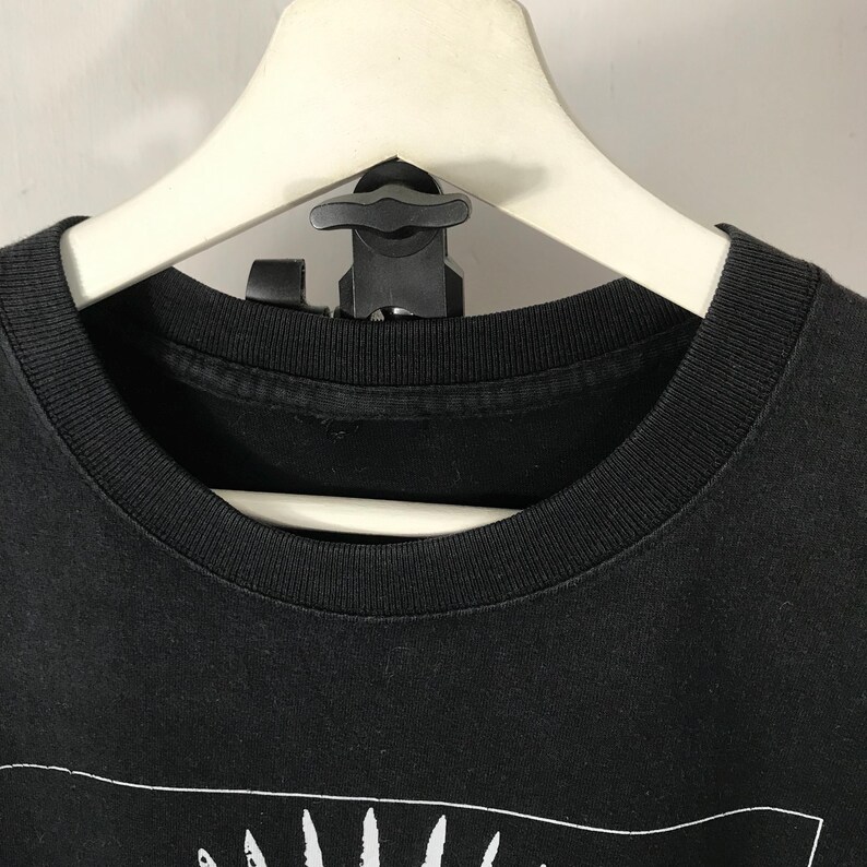 Może przedstawiać: Czarny t-shirt z okrągłym dekoltem i białym wzorem graficznym na dole. Koszulka wisi na białym wieszaku z czarnym zaciskiem. T-shirt jest wykonany z miękkiego materiału.