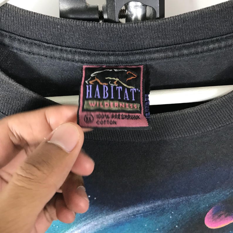 Puede incluir: Camiseta gris oscuro con un gr&aacute;fico colorido con tem&aacute;tica espacial. La camiseta presenta una etiqueta con las palabras "HABITAT WILDERNESS" y una silueta de oso. Hecha de 100% algod&oacute;n pre-encogido.