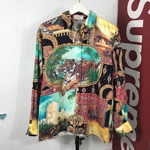 Puede incluir: Camisa abotonada de manga larga con un estampado vibrante y colorido que presenta un tigre, elementos arquitectónicos y patrones ornamentados. La camisa de cuello está colgada en una percha blanca.
