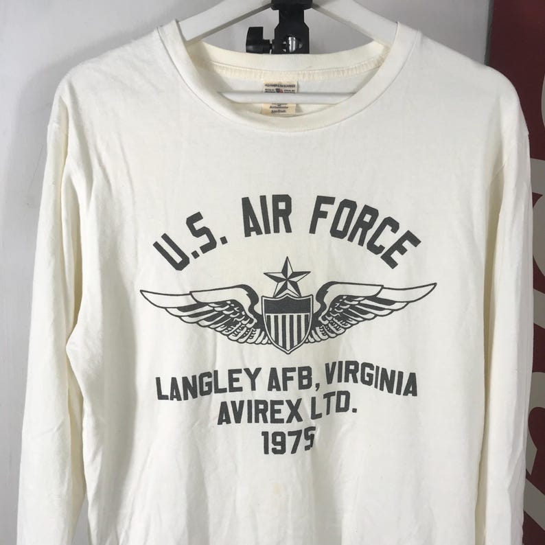 May include: Off-white long-sleeved shirt featuring the text "U.S. AIR FORCE" above a graphic of wings, a star, and a shield. Below the graphic, the text reads "LANGLEY AFB, VIRGINIA AVIREX LTD. 1975".