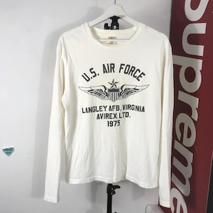 May include: Cream-coloured long-sleeved shirt featuring the text "U.S. AIR FORCE" above a graphic of wings and a star. Below the graphic is "LANGLEY AFB, VIRGINIA AVIREX LTD. 1975". The shirt is hanging on a white hanger.