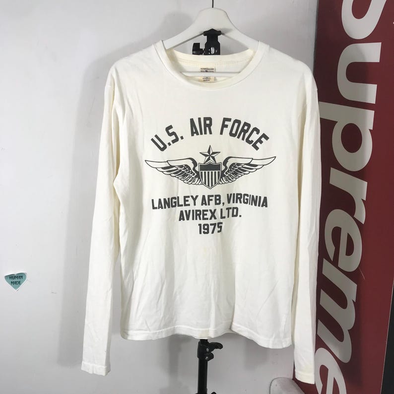 May include: Cream-coloured long-sleeved shirt featuring the text "U.S. AIR FORCE" above a graphic of wings and a star. Below the graphic is "LANGLEY AFB, VIRGINIA AVIREX LTD. 1975". The shirt is hanging on a white hanger.