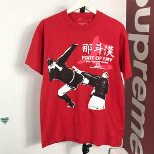 Puede incluir: Camiseta roja con un gráfico en blanco y negro de un jugador de fútbol pateando un balón. La camiseta tiene el texto "FOOT OF FURY" y caracteres chinos. La camiseta está colgada en una percha blanca.