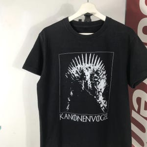 Może przedstawiać: Czarny t-shirt z białą grafiką przypominającą koronę w prostokącie. Tekst "KANONENVOGEL" jest wydrukowany poniżej na biało. Koszulka wisi na białym wieszaku.