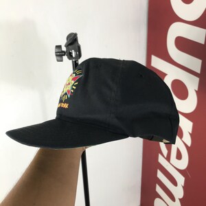 May include: Black baseball cap with a colorful graphic on the front. The graphic includes a starburst design with yellow, red, and green accents. The text "NEW YORK" is visible on the cap.