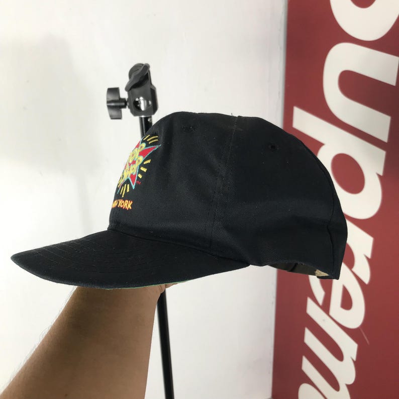 May include: Black baseball cap with a colorful graphic on the front. The graphic includes a starburst design with yellow, red, and green accents. The text "NEW YORK" is visible on the cap.