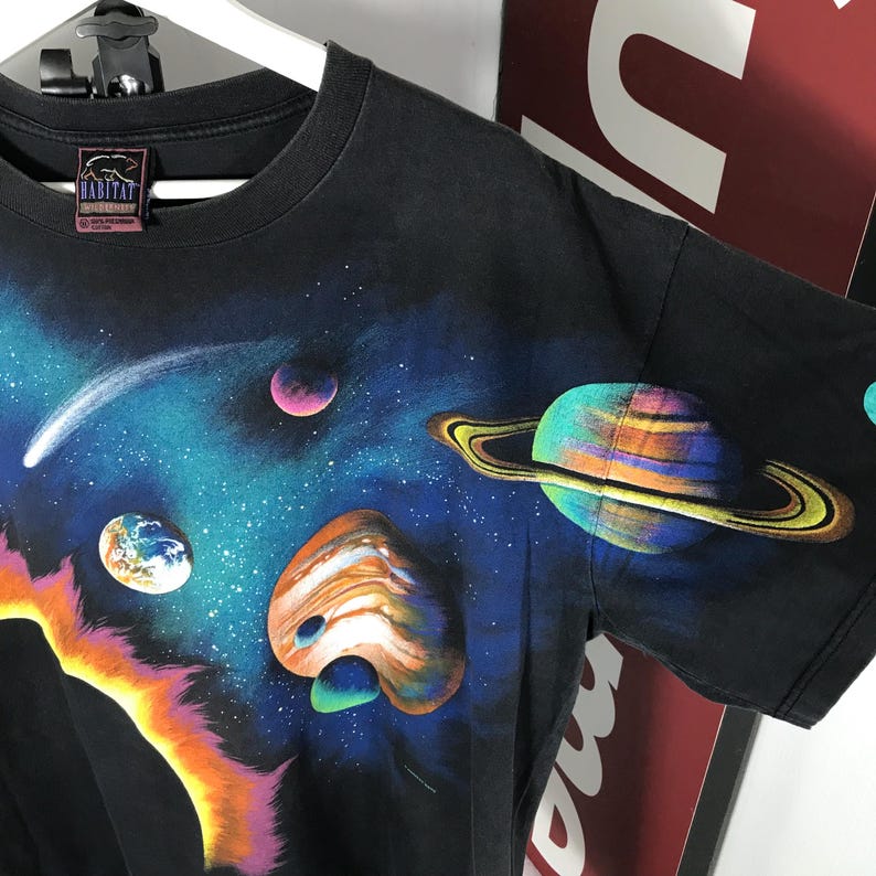 Puede incluir: Camiseta negra con un vibrante dise&ntilde;o tem&aacute;tico espacial. El dise&ntilde;o incluye planetas, estrellas y un cometa sobre un fondo azul oscuro y p&uacute;rpura. La camiseta tiene cuello redondo y mangas cortas.