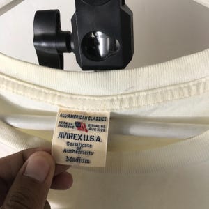 May include: Cream-coloured t-shirt with a tag from AVIREX U.S.A. The tag reads "ALL-AMERICAN CLASSICS" and includes the text "Certificate of Authenticity" and "Medium". The shirt is held by a person.