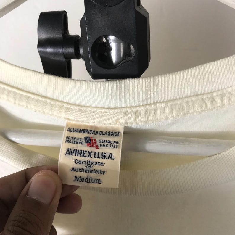 May include: Cream-coloured t-shirt with a tag from AVIREX U.S.A. The tag reads "ALL-AMERICAN CLASSICS" and includes the text "Certificate of Authenticity" and "Medium". The shirt is held by a person.