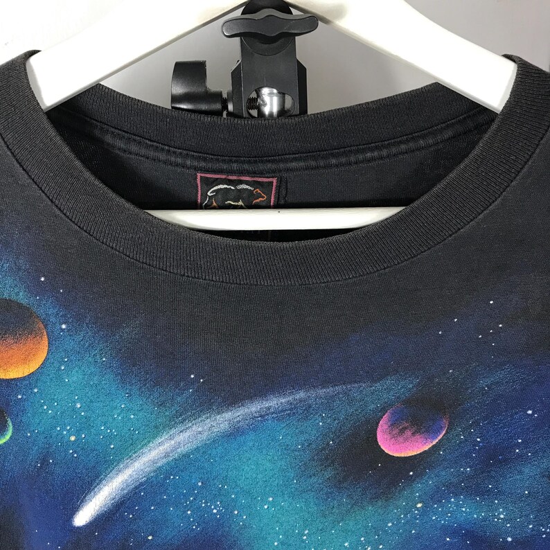 Puede incluir: Camiseta negra con un dise&ntilde;o vibrante de tem&aacute;tica espacial que muestra planetas, estrellas y un cometa sobre un fondo azul y turquesa. La camiseta tiene cuello redondo y una etiqueta con un logo de oso.