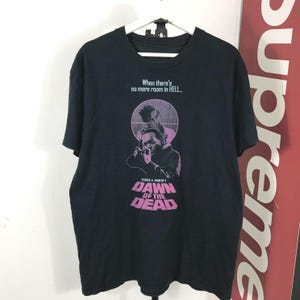 May include: A black short-sleeve t-shirt featuring a graphic print from the film Dawn of the Dead. The design includes the text When there's no more room in HELL and George A. Romero's DAWN OF THE DEAD in pink and white ink on the chest.