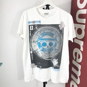 Puede incluir: Camiseta blanca con un diseño gráfico de la serie de anime "One Piece". El diseño incluye el logotipo de la serie y un emblema de calavera y huesos cruzados en azul y blanco. La camiseta tiene mangas cortas y cuello redondo.
