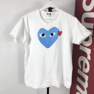 May include: White t-shirt featuring a large blue heart with two eyes and a small red heart on the chest. The shirt has a crew neck and short sleeves. The word "PLAY" is printed on the tag.