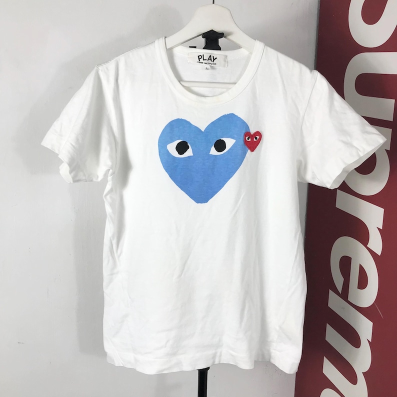 May include: White t-shirt featuring a large blue heart with two eyes and a small red heart on the chest. The shirt has a crew neck and short sleeves. The word "PLAY" is printed on the tag.