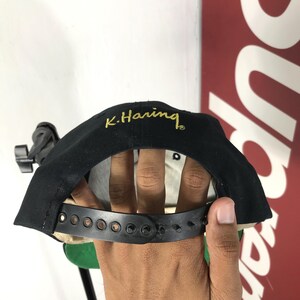 May include: Black baseball cap with the embroidered text "K. Haring" in gold. The cap features a black adjustable strap and a curved brim. The interior lining is tan. A classic style hat.