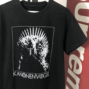 Może przedstawiać: Czarny t-shirt z białym nadrukiem przedstawiającym tron i twarz kobiety oraz napisem "KANONENVOGEL" poniżej. Koszulka ma okrągły dekolt i kr&oacute;tkie rękawy. Projekt jest wyśrodkowany z przodu.
